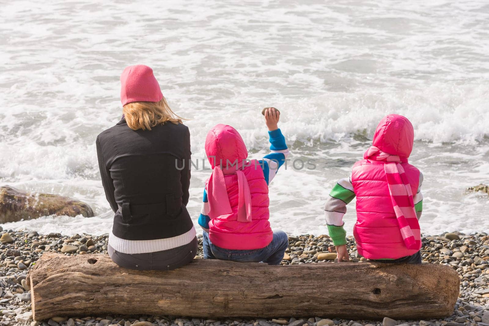 Two girls and woman sitting on a log on the beach and throwing stones into the water