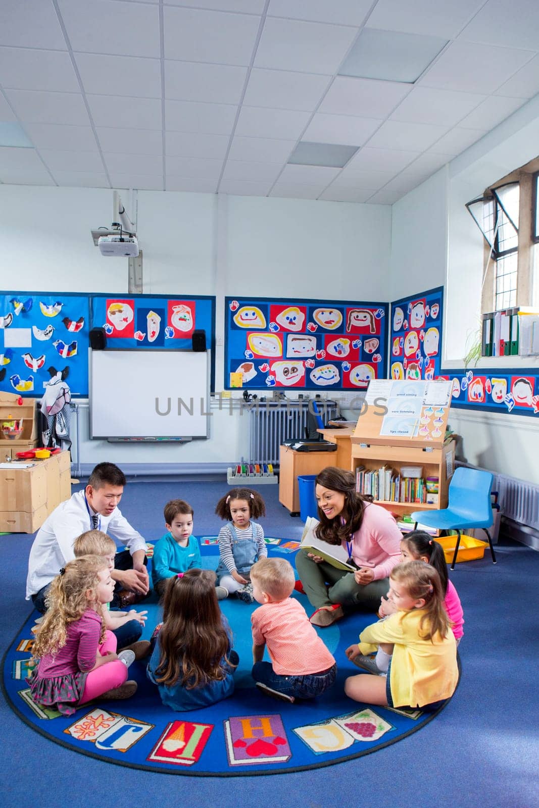 Group of nursery children sitting on the floor in their classroom with their teachers. The female teacher is reading from a book.
