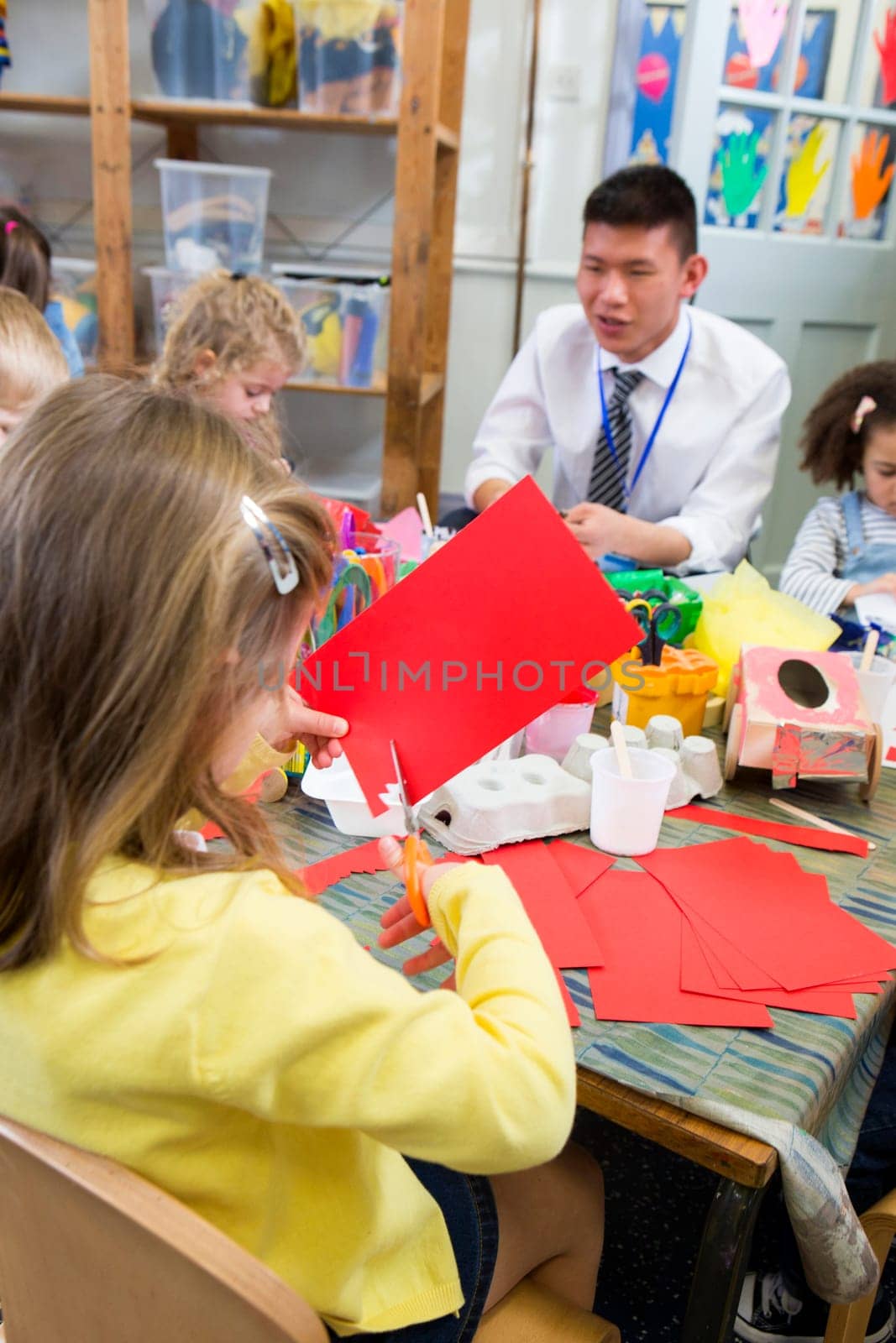 Little girl using scissors to cut shapes from a red piece of paper. She is in a nursery class with other students and a teacher.