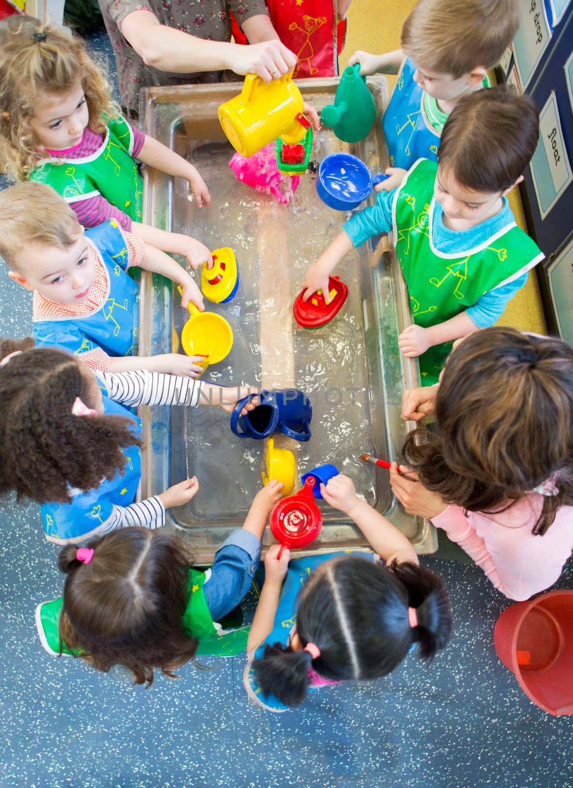 Overhead view of a class of nursery children playing at a water table. They are all wearing aprons and using plastic toys.