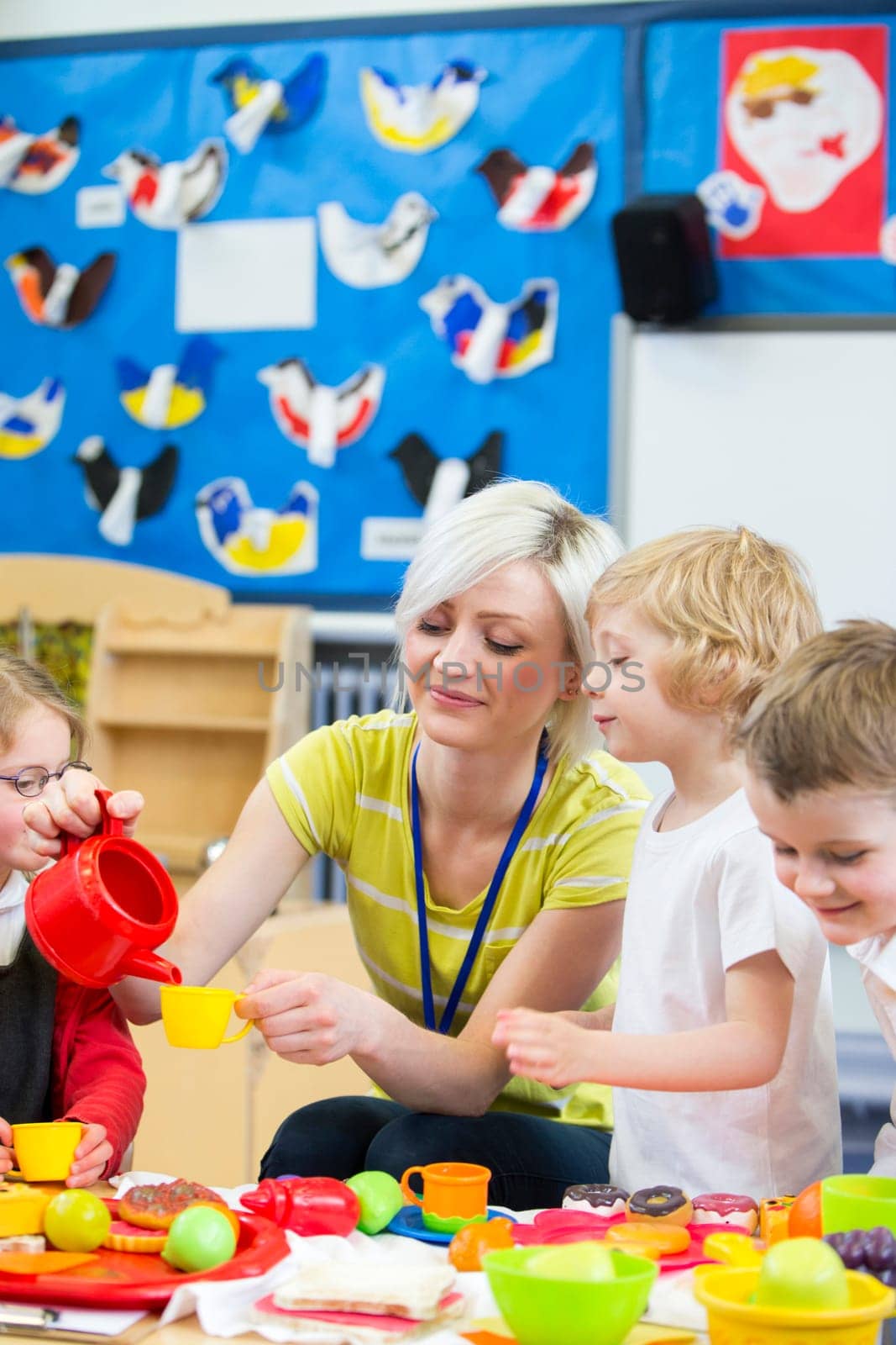 Nursery teacher playing kitchen roleplay with her students in the classroom. she is pouring a pretend cup of tea for a little boy.