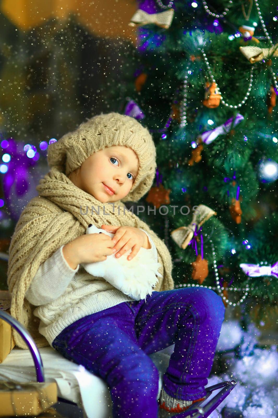the girl in lilac jeans, in a knitted cap and with a knitted scarf sits on a sledge near a Christmas fir-tree with a toy and it is snowing