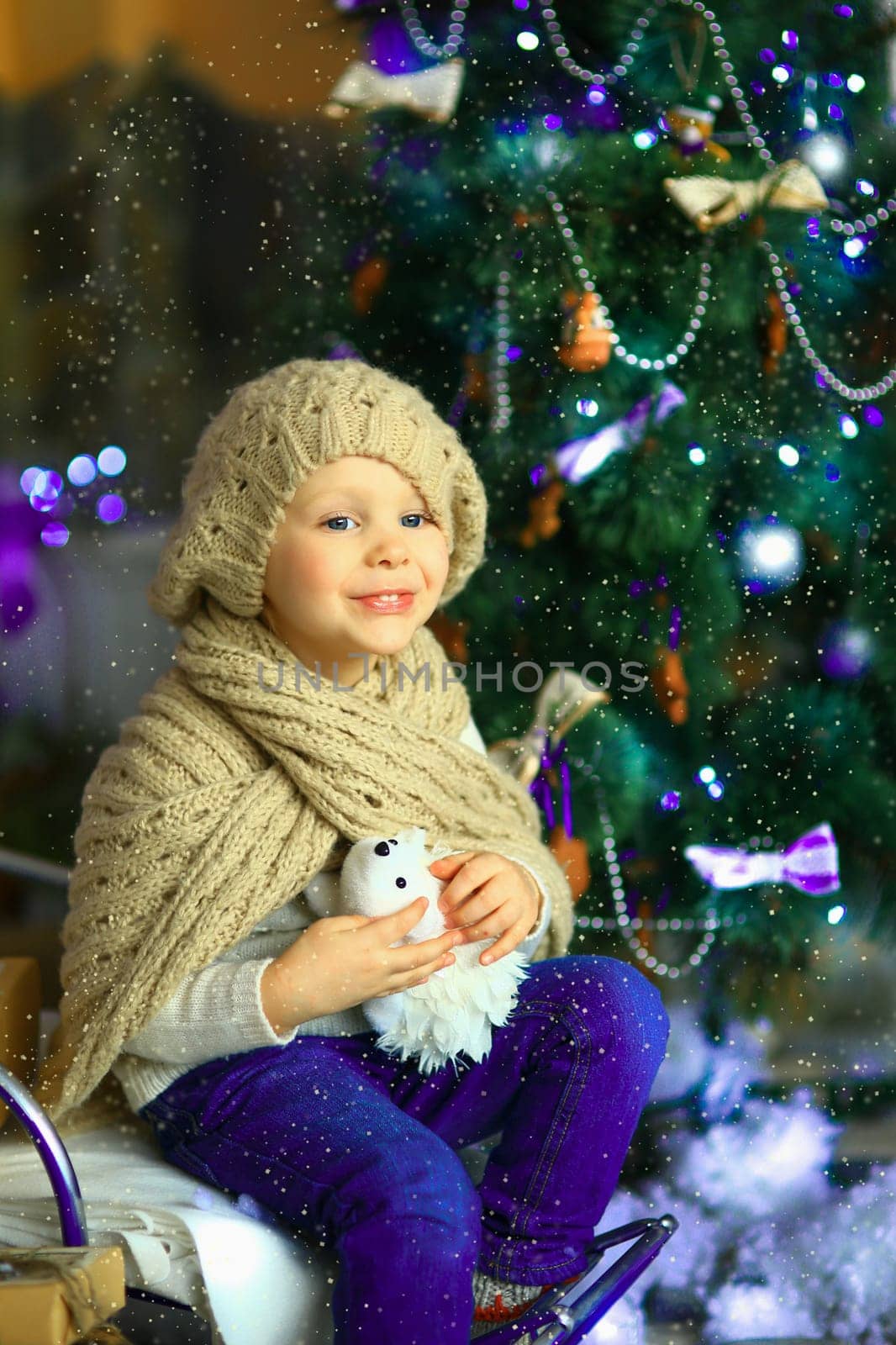 the girl in lilac jeans, in a knitted cap and with a knitted scarf sits on a sledge near a Christmas fir-tree with a toy and it is snowing