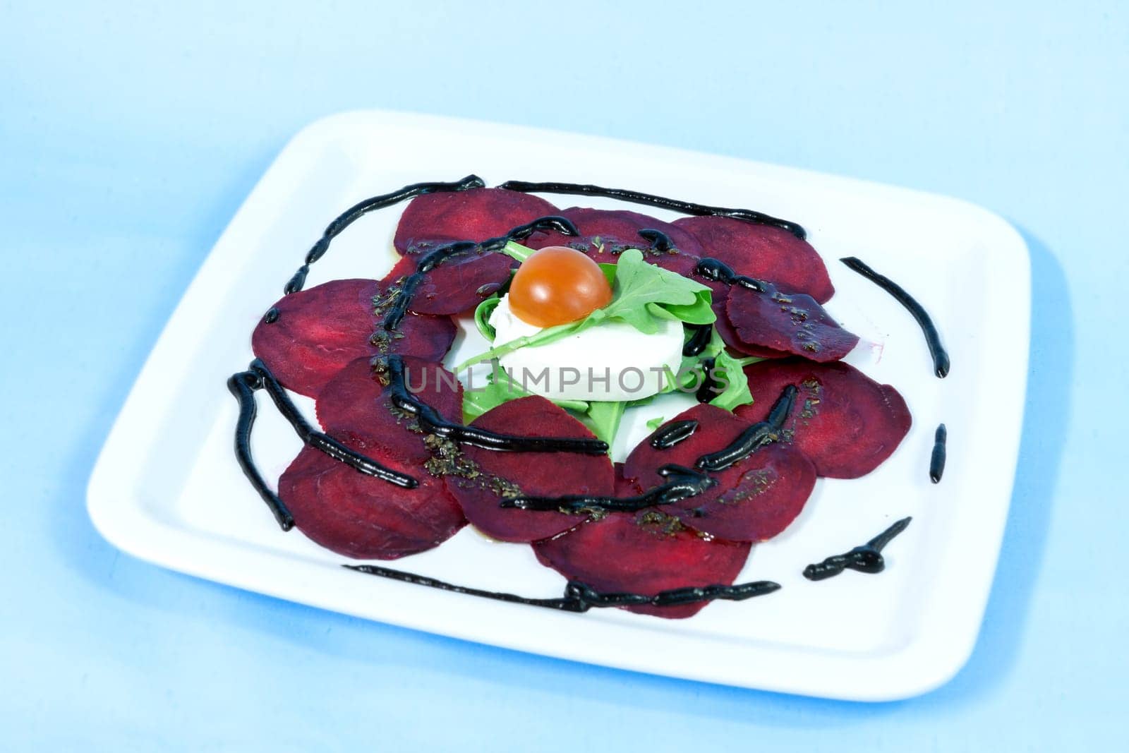 Modern food on a white plate and blue background - Thin slices of beetroot with goat cheese and balsamic reduction