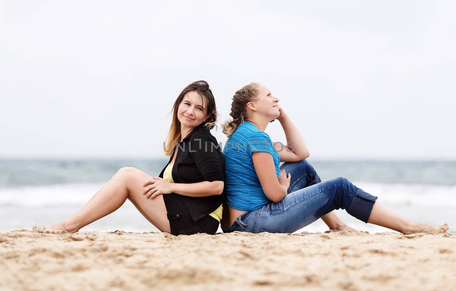 Two pretty young beautiful smiling women sitting with his back to each other and resting on the sand on the seashore. Two girls resting on the beach. Two girls outdoors. Selective focus on the models.