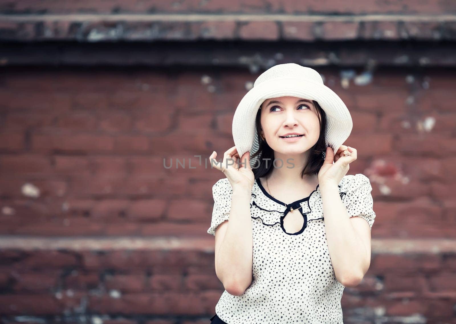 Pretty young smiling woman in a white blouse and hat posing on old vintage brick wall background. Girl holds the edges of the hat by hand. Toned photo with copy space. Vintage style photo.