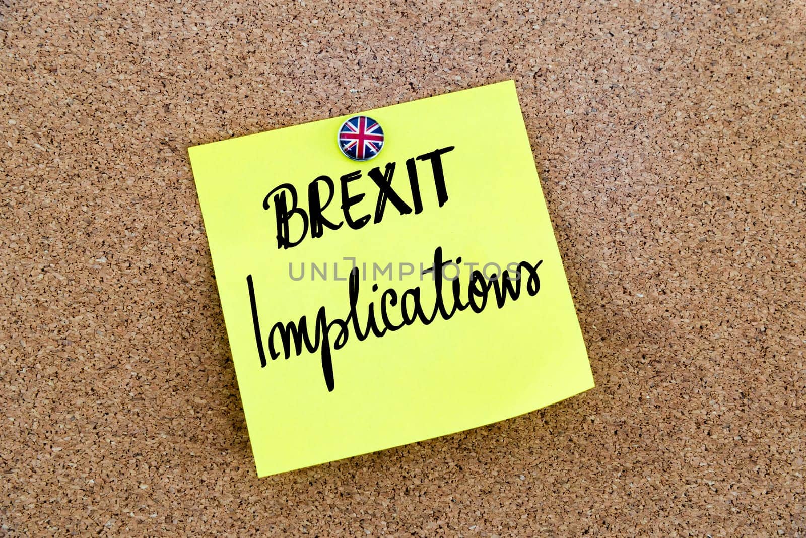 Yellow paper note pinned on cork board with Great Britain flag thumbtack, written text BREXIT IMPLICATIONS, United Kingdom exit from European Union