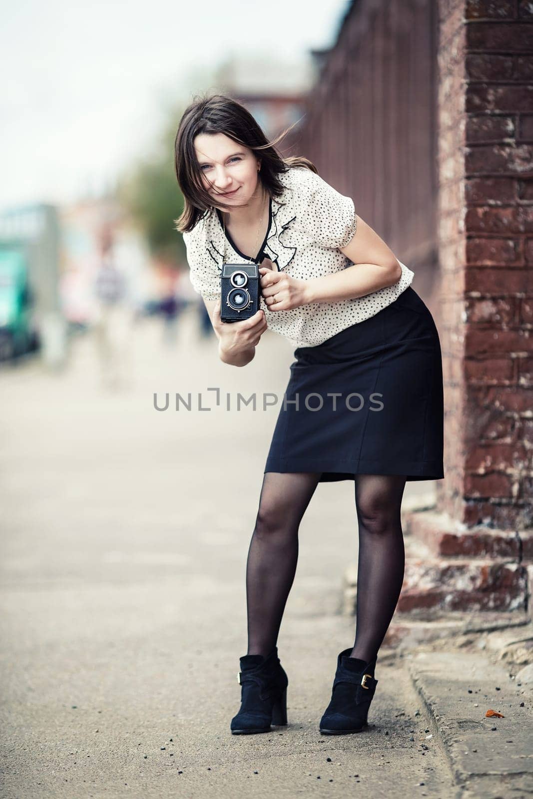 Girl holding retro camera and taking photo. Pretty young woman photographer with old vintage camera outdoors. Retro style photo. Selective focus on model. Vertical shot. Vintage style photo.