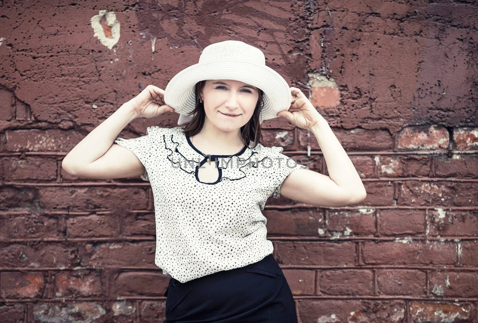 Pretty young woman in white blouse and hat posing against brick wall background. Woman holds the edges of the hat by hands. Toned photo with copy space. Vintage style photo.