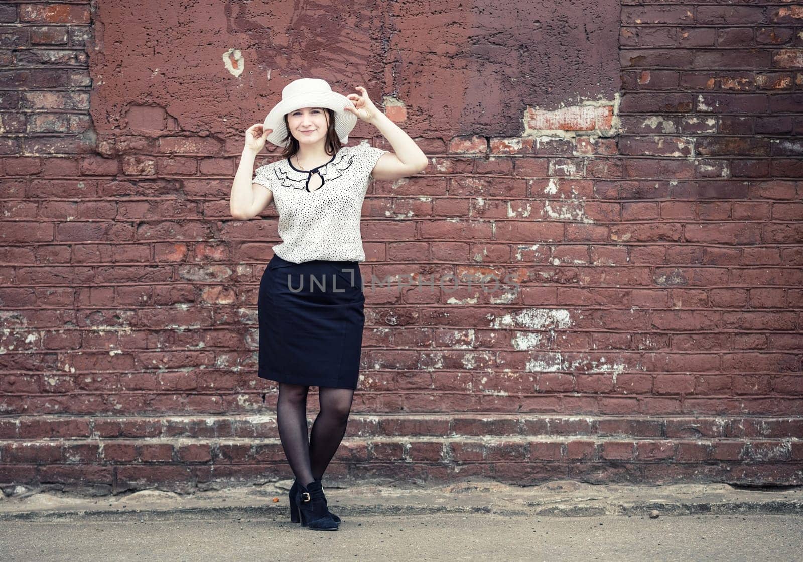 Pretty young woman in white hat, blouse and black skirt, standing against brick wall background. Girl holds the edges of the hat by hands. Toned photo with copy space. Vintage style photo.