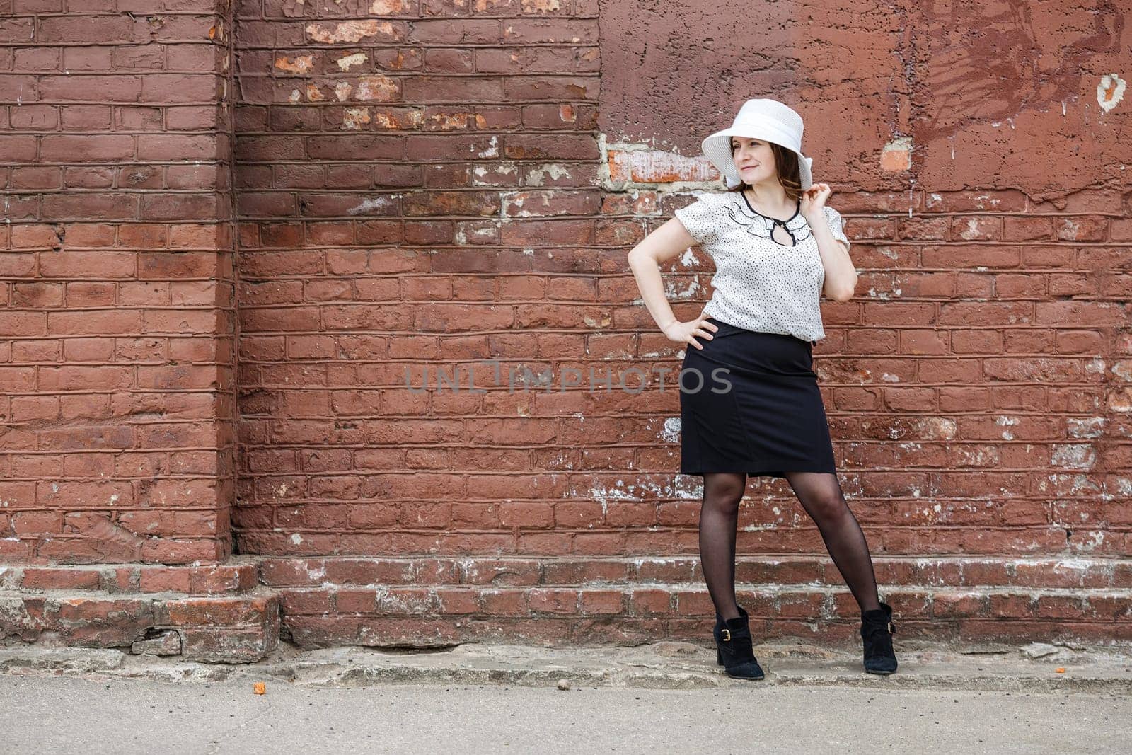 Woman in a white hat, blouse and black skirt, posing against the backdrop of an old vintage brown brick wall. Female portrait in a retro style.