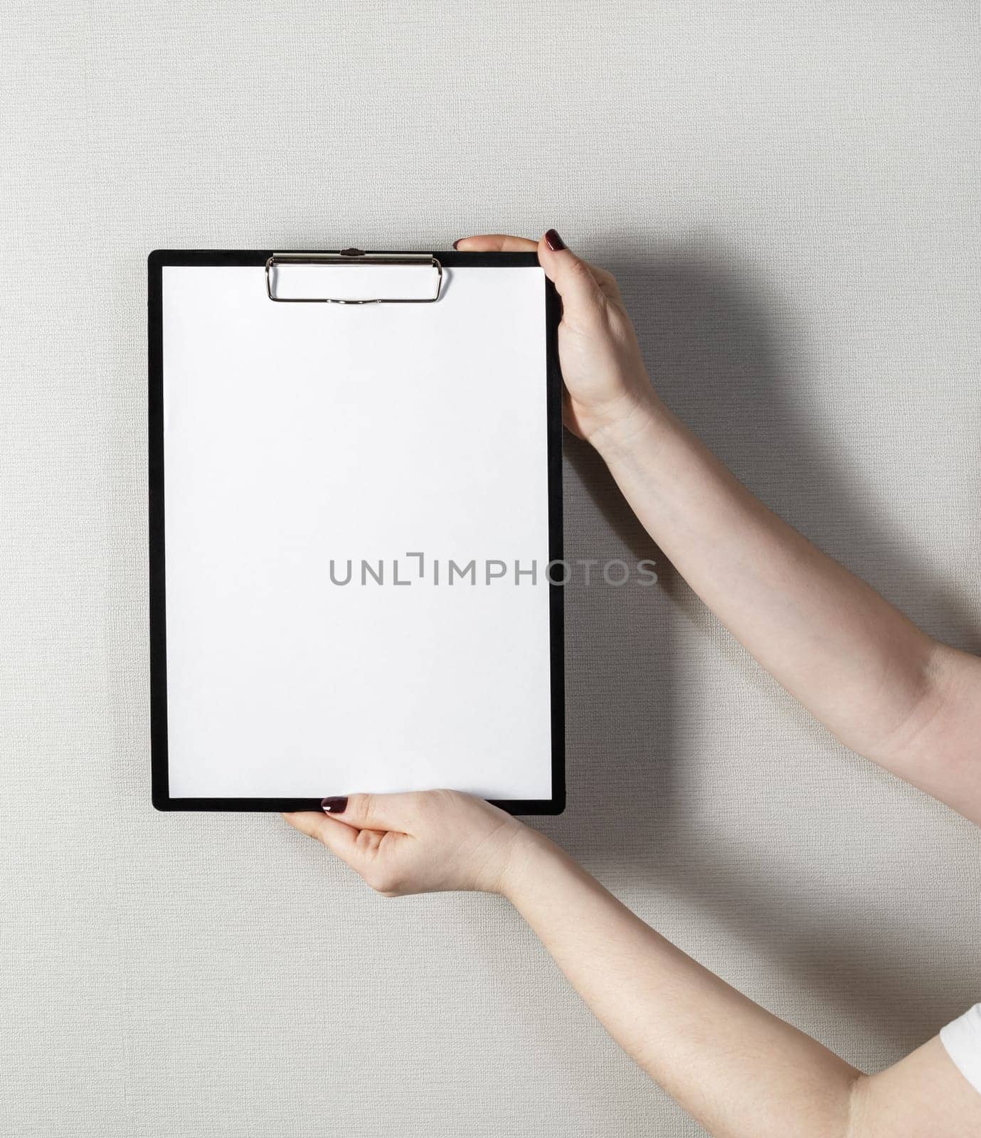 Clipboard with a blank sheet of paper in female hands with a deep shadow on the background of gray wall. Mock-up for design presentations and portfolios.