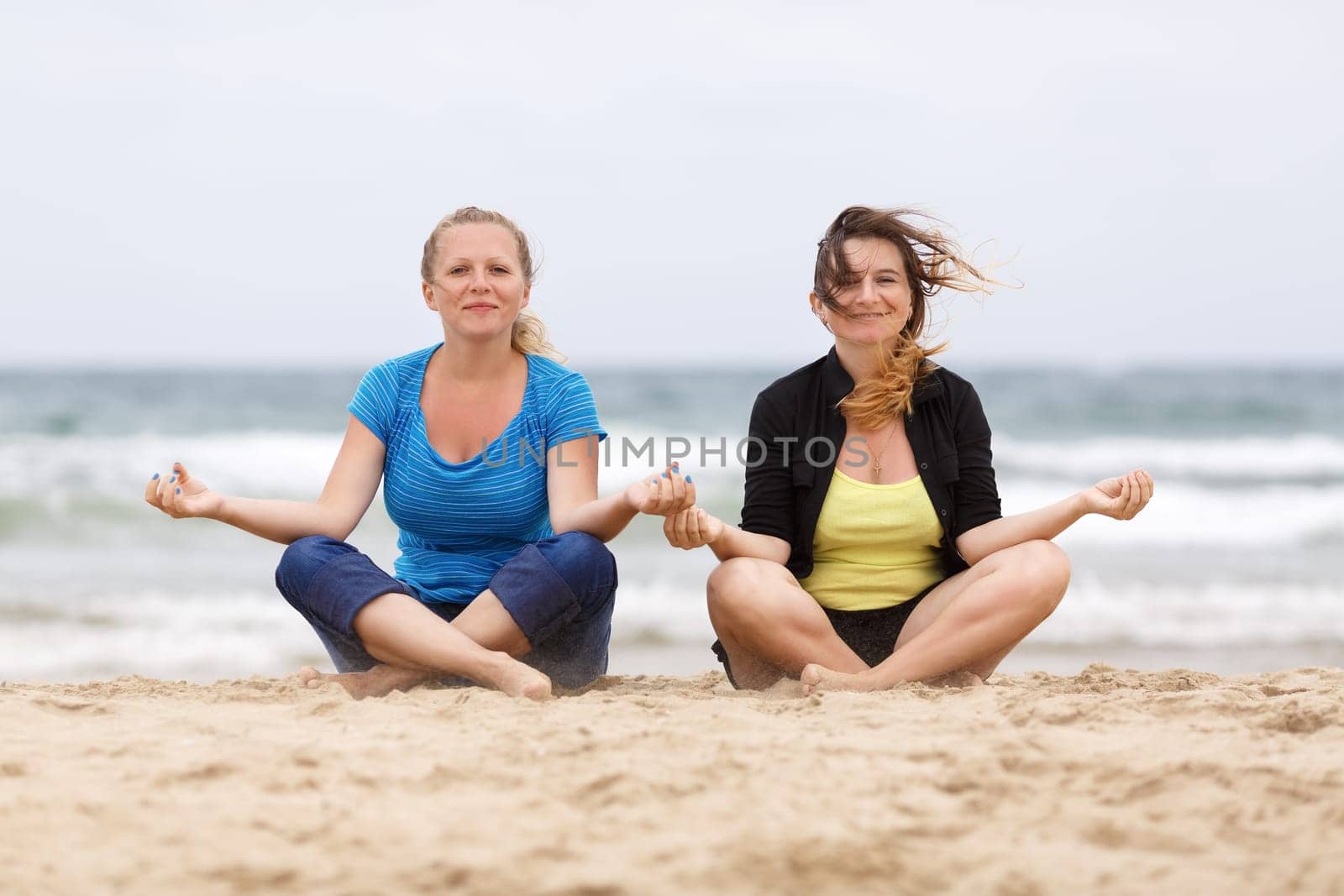 Two pretty young smiling woman sitting in lotus pose on the sand on the beach and relax. Shallow depth of field. Focus on models.
