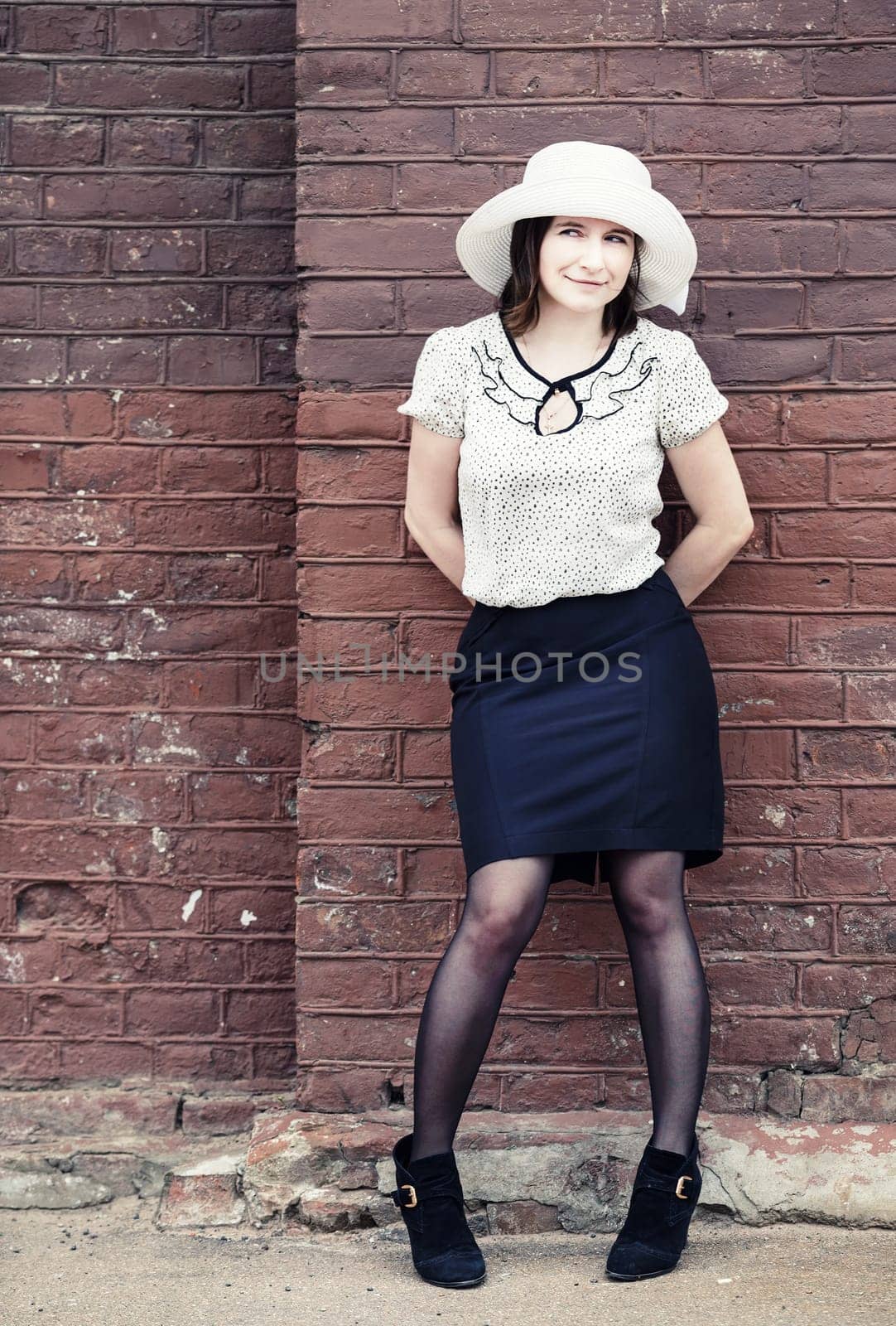 Pretty young woman in a white hat, blouse and black skirt, posing outdoor against brick wall background. Toned photo with copy space. Vintage style photo.