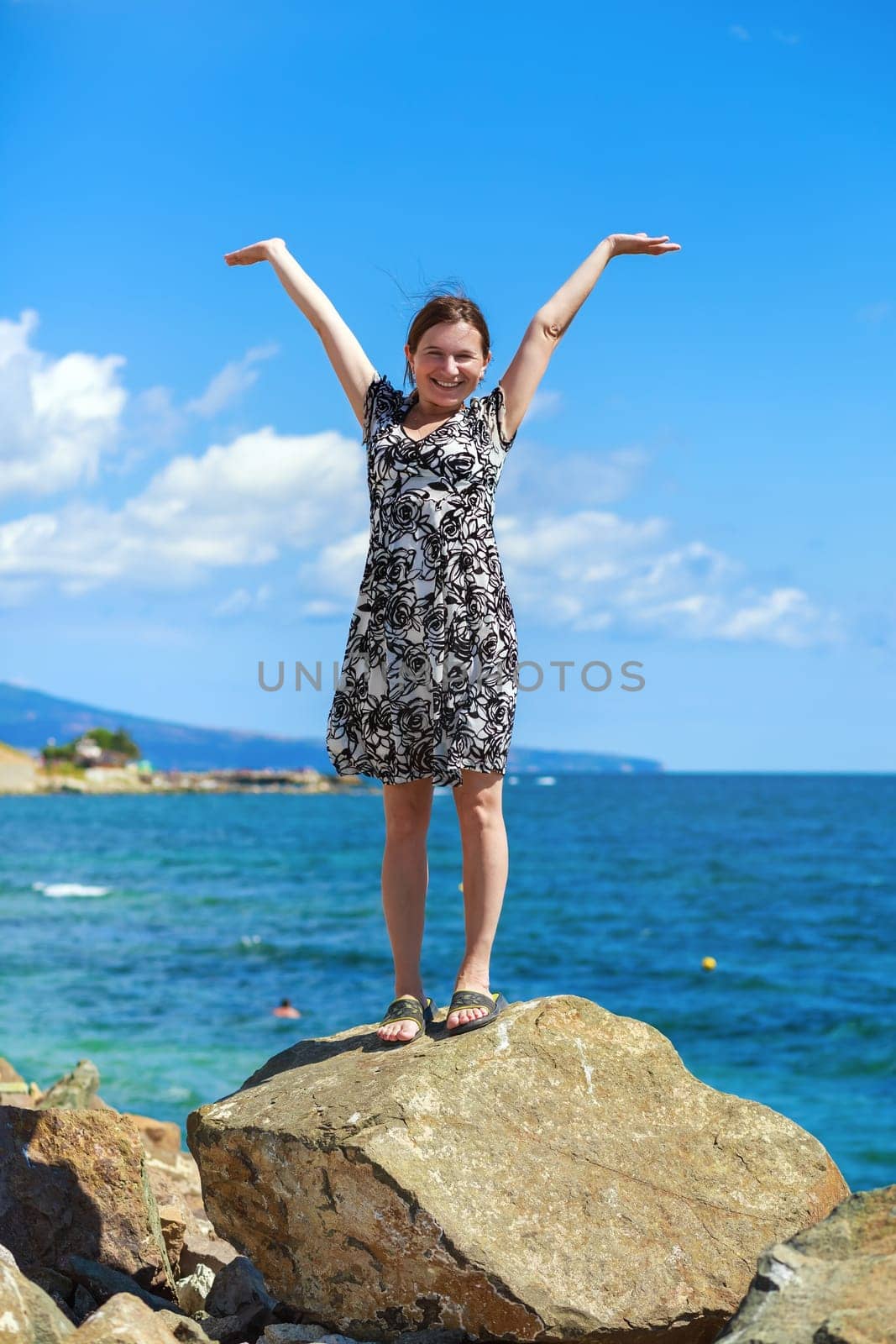 Pretty young woman standing on a stone and posing with raised hands against a background of sea and blue sky. Selective focus on the model.