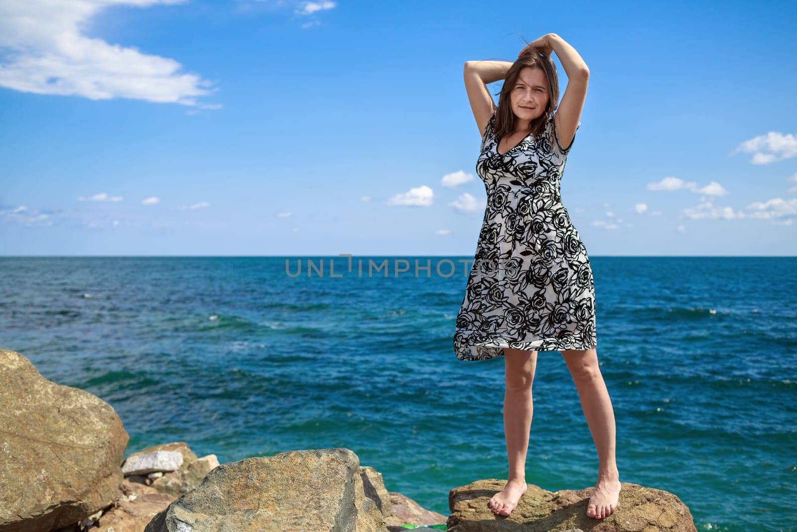 Pretty young woman standing on a stone and posing with his hands behind his head against a background of sea and blue sky. Selective focus on the model.