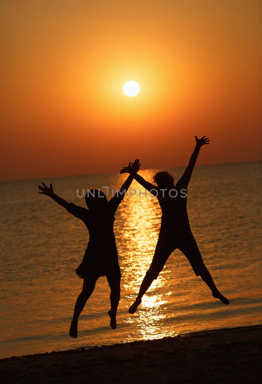 Two girls jumping on a background of the rising sun over the sea. Focus on models. Shallow depth of field. Toned image.