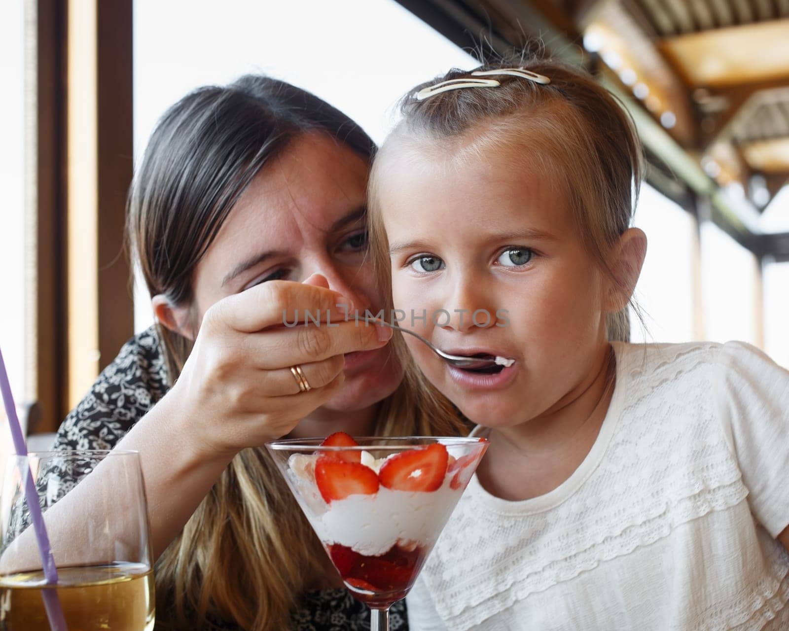 Mother feeds his daughter delicious sweet ice cream with strawberries in a restaurant. Shallow depth of field. Focus on the child's face.