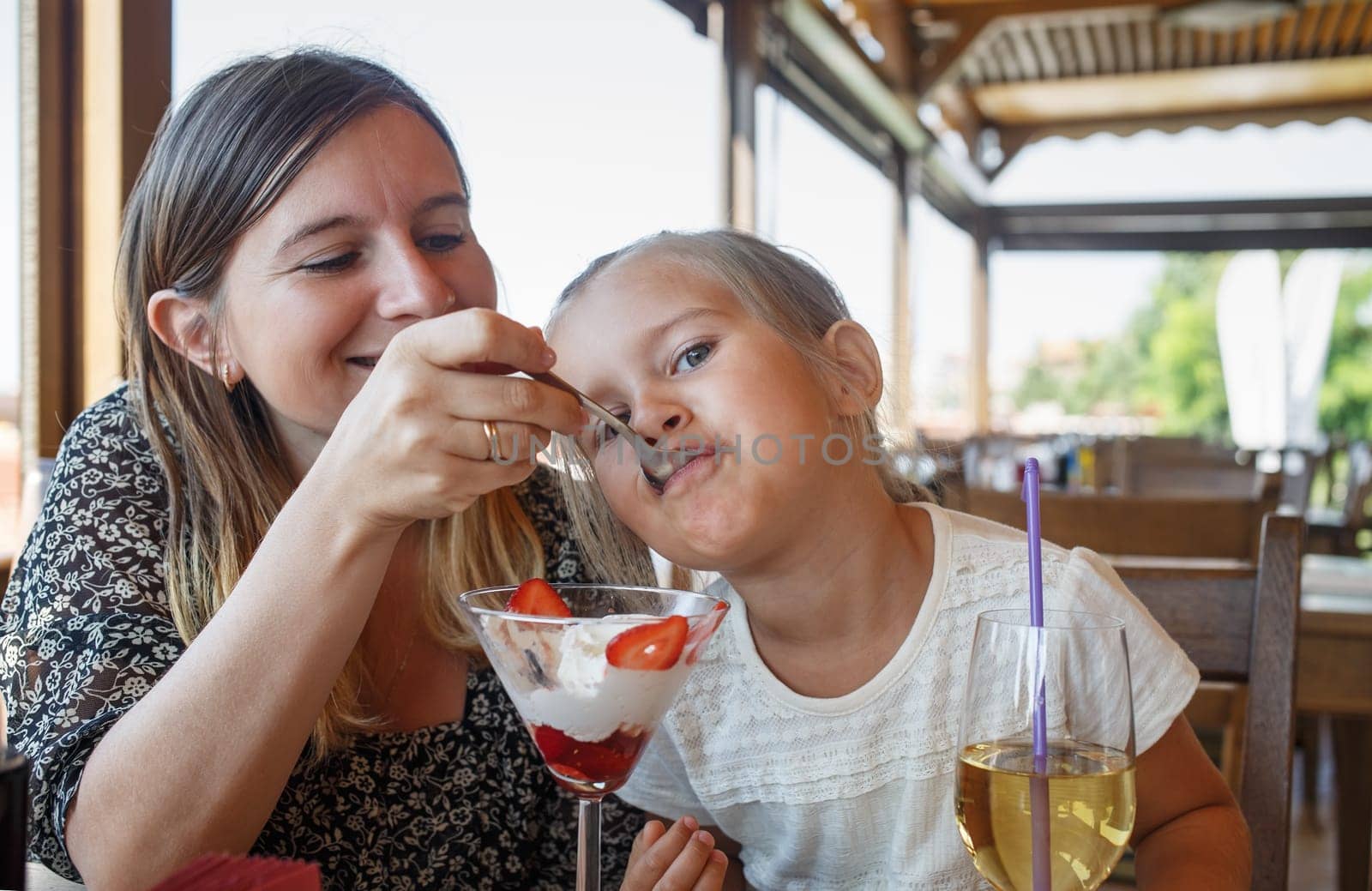Mum feeds the daughter delicious sweet ice cream with strawberries in a restaurant. Shallow depth of field. Focus on the child's face.