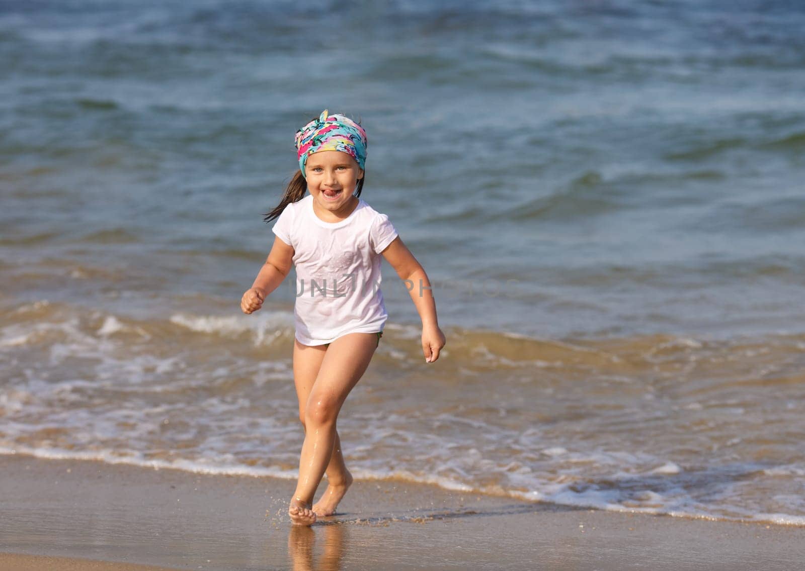 Smiling child with his tongue hanging out in a white t-shirt runs the water's edge on the beach on a clear sunny day. Shallow depth of field. Focus on the model's face.