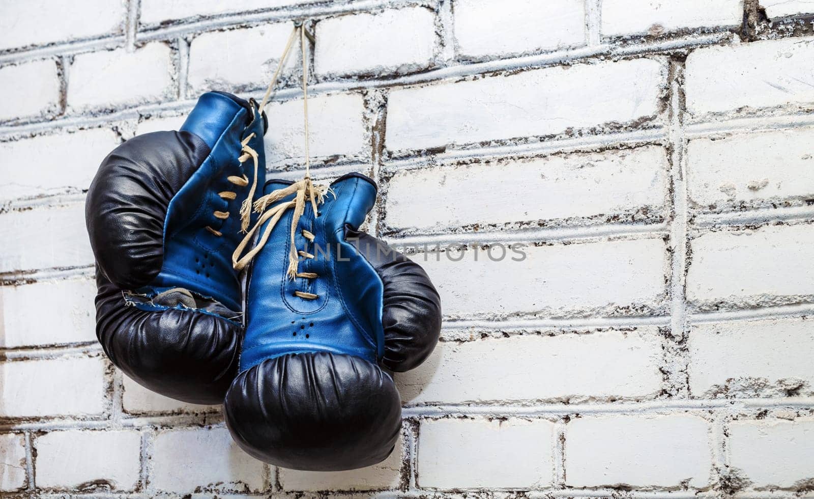 A pair of old blue and black boxing gloves hanging on white brick wall background.