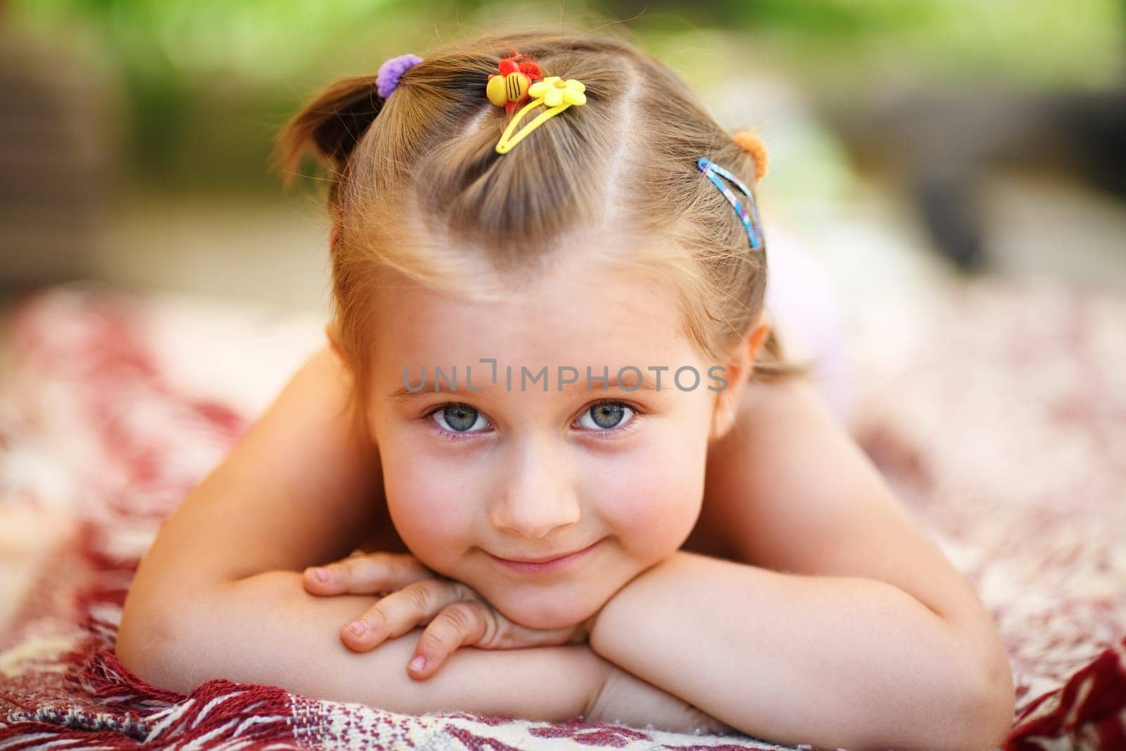 Sweet happy little baby girl lying on a blanket in the park. Close-up of child resting her head on her hands and looking into the camera. Shallow depth of field. Selective focus.