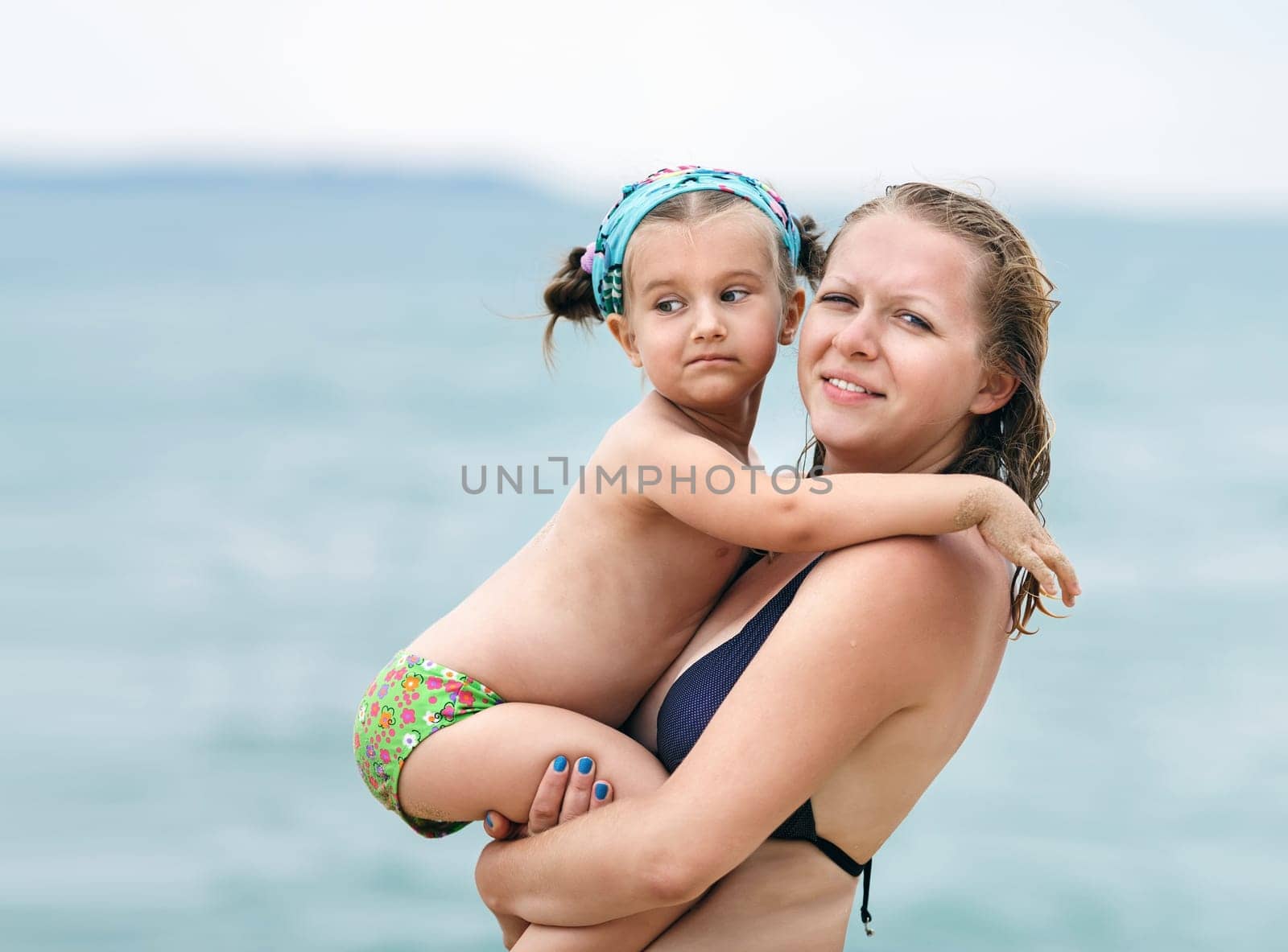 Hugging mother and daughter. Mom keeps little daughter in her arms on  blurred background of the sea. Selective focus on the models.