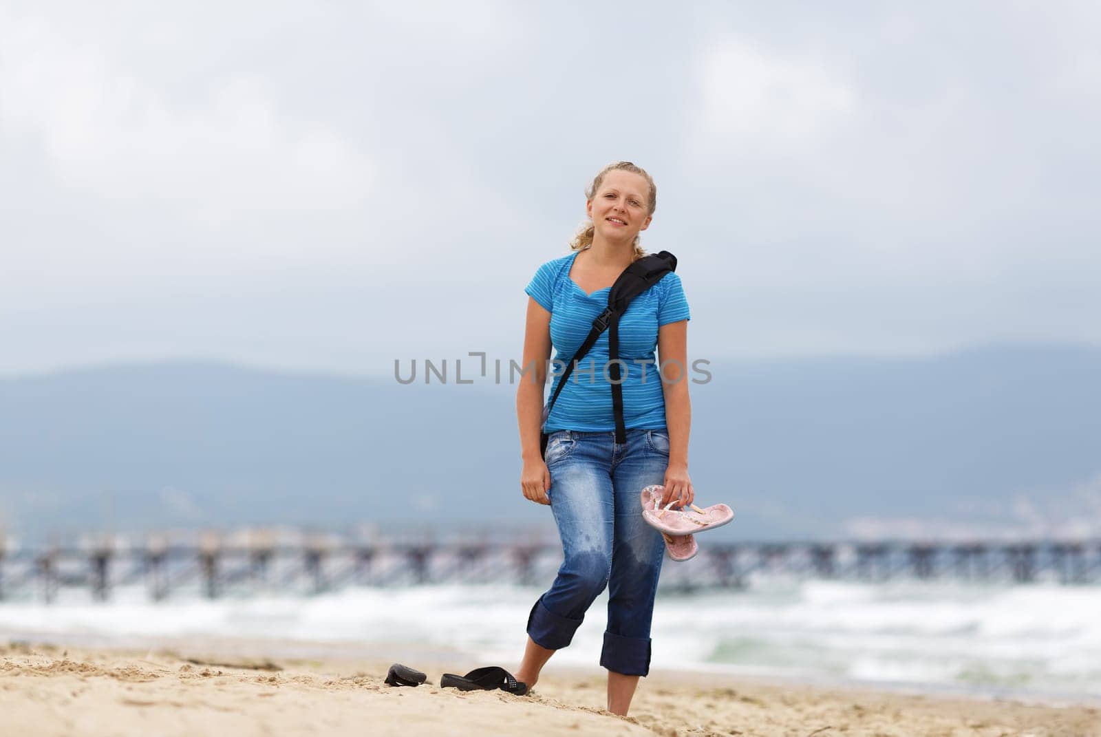 Young smiling blonde woman barefoot in blue t-shirt and jeans with a backpack standing on the beach against the sea.  Shallow depth of field. Focus on model.