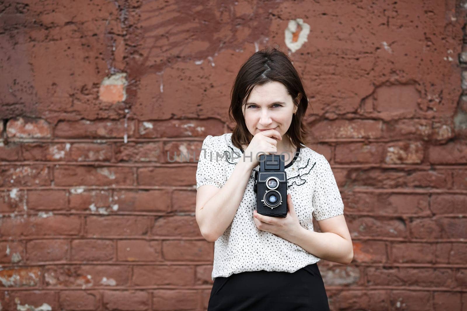 Pretty young woman photographer with retro photocamera in hand. Model is thinking and looking at camera. Old brown vintage grunge wall in the background.