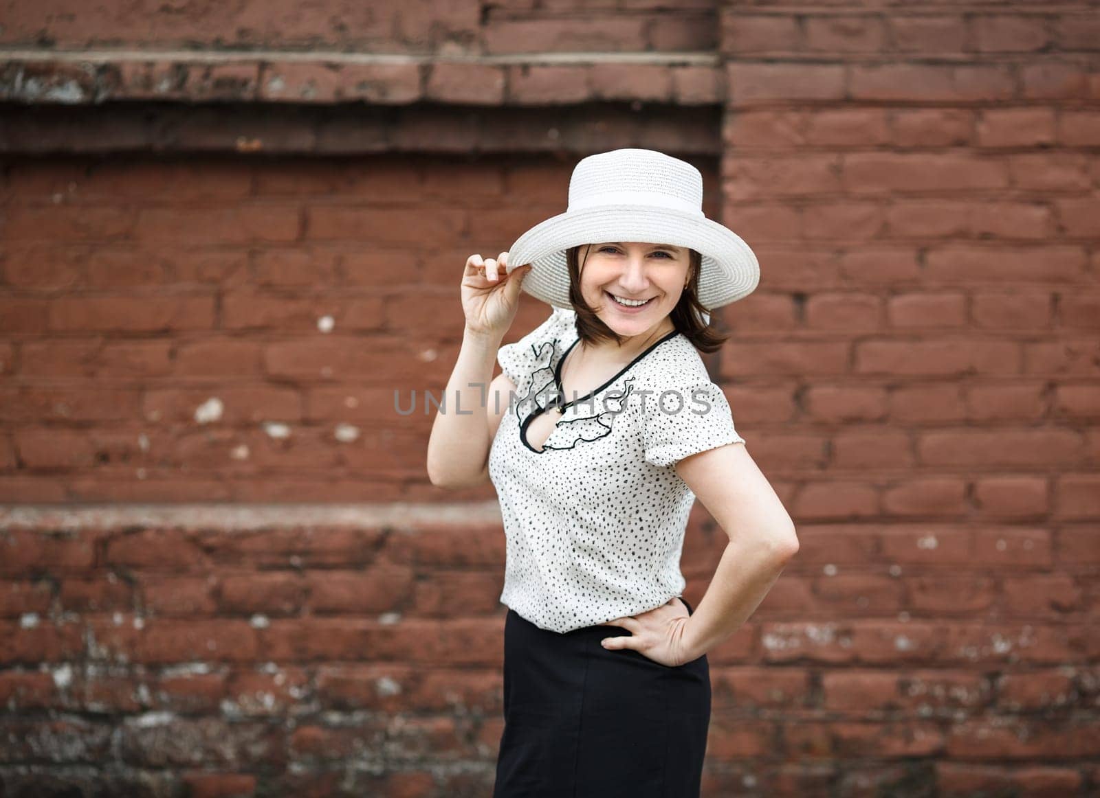 A smiling young woman in a white blouse and a hat posing against the backdrop of an old vintage brown brick wall. Female hand holds a hat.  Photo in retro style. Selective focus on model.