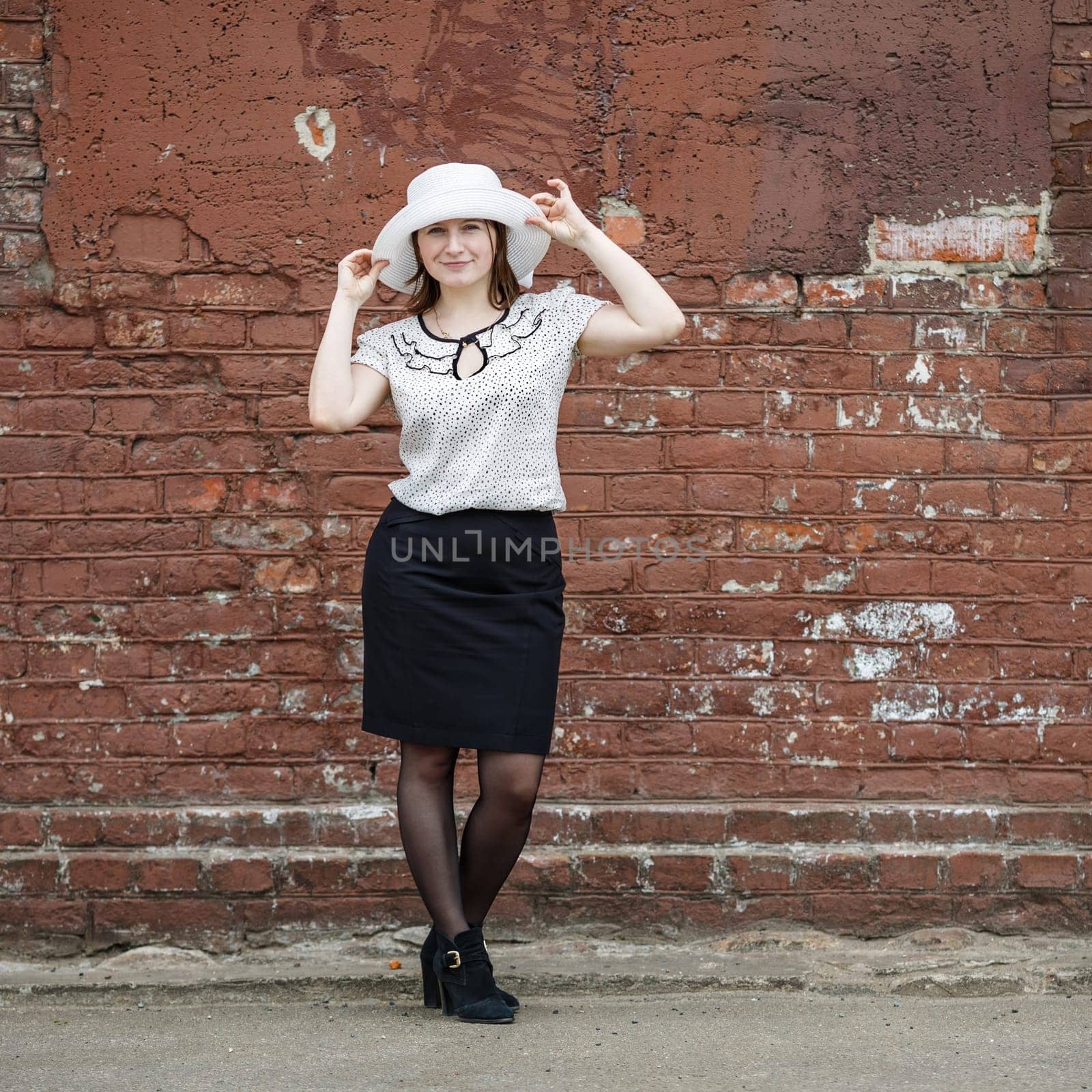 Photo of a woman in a white hat, blouse and black skirt, standing against the backdrop of an old vintage brown brick wall. Model holds the edges of the hat by hand. Portrait of a woman in a retro style.