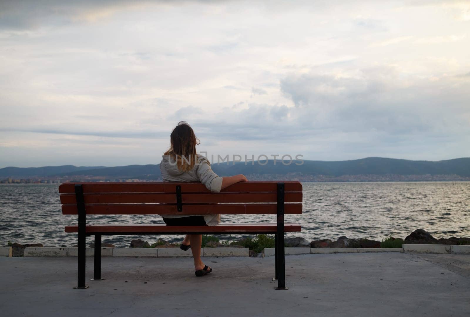 A woman sitting on a bench in the early morning and admire views of the bay and mountains. View from the back. Shallow depth of field. Focus on model.