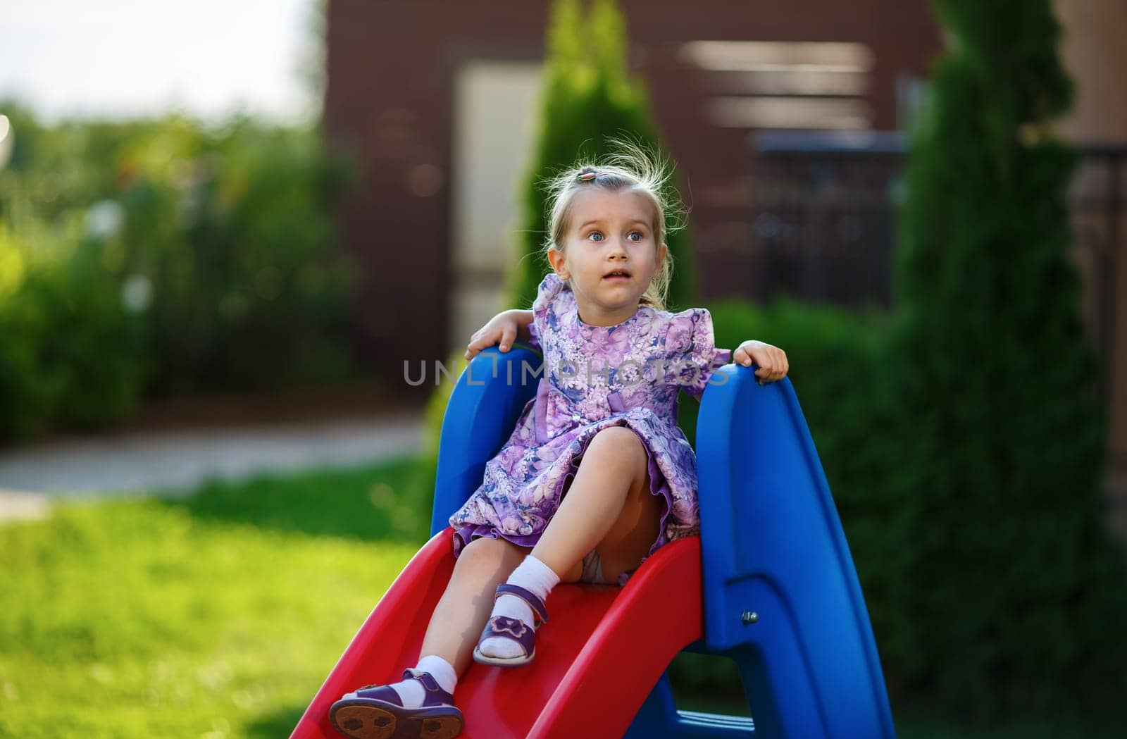 A child in a smart dress and sandals sitting on slide and dreamily looking up. Cute little girl. Warm sunny summer day. Shallow depth of field. Selective focus on the model's face.
