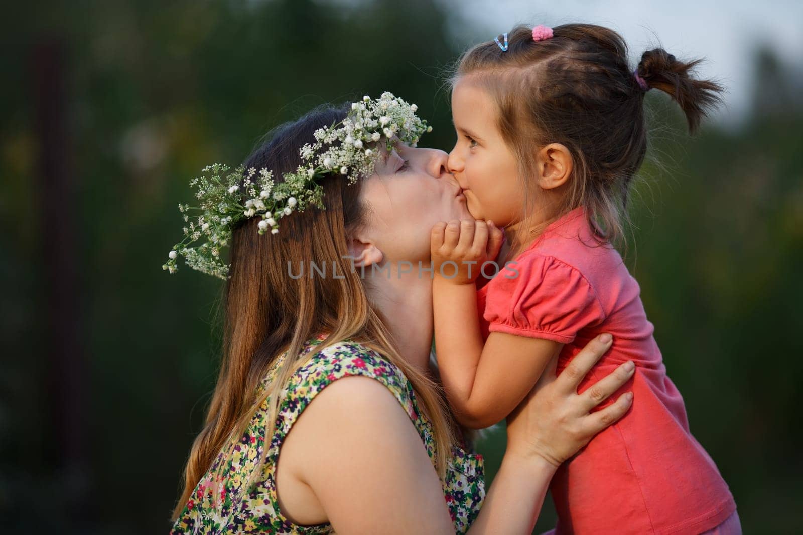 Happy mother wearing a wreath of wildflowers and daughter kissing. Sunset lighting. Shallow depth of field. Focus on models.