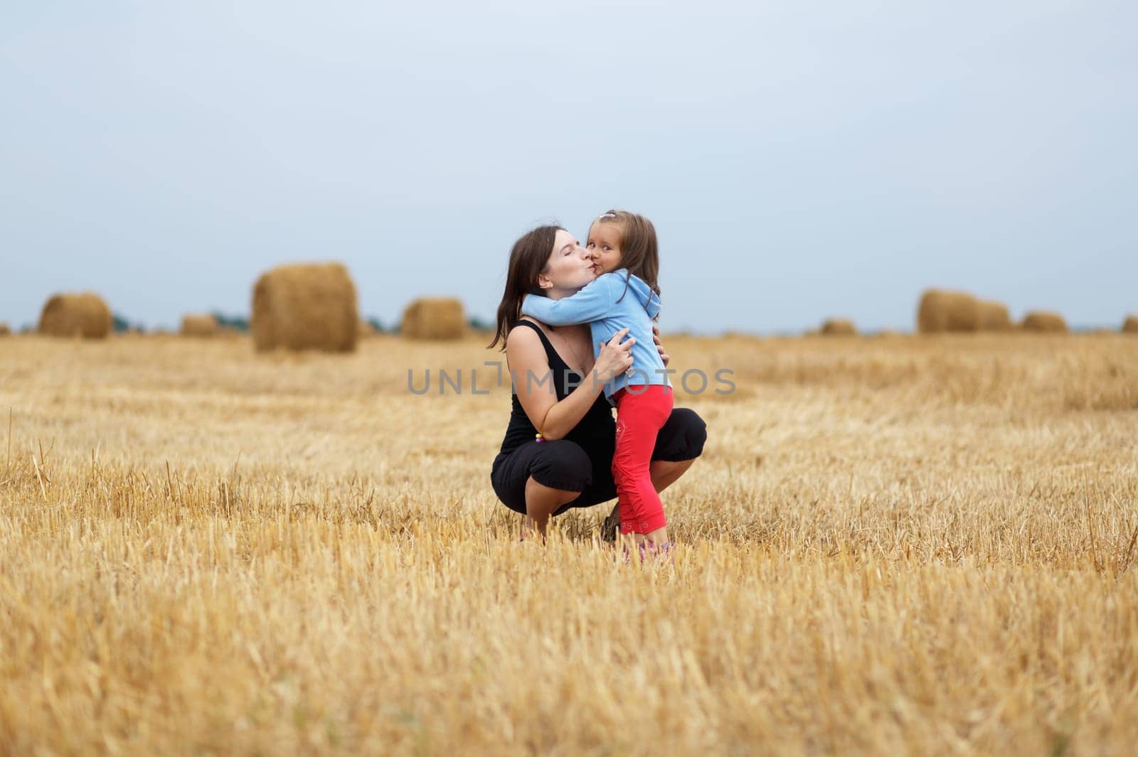 Mother and daughter hugging in the middle sloping field on straw bales background. Shallow depth of field. Selective focus on the models.