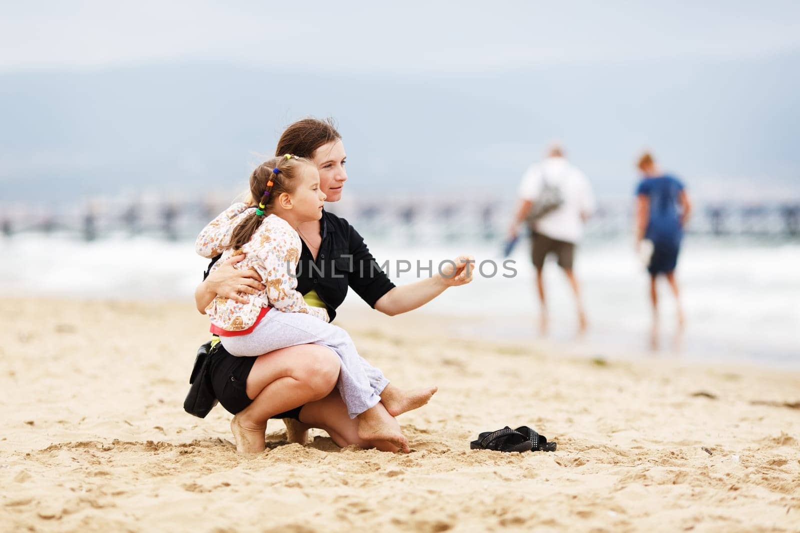 Pretty young mother sitting on the beach and holding his little daughter on his knee. Shallow depth of field. Selective focus.