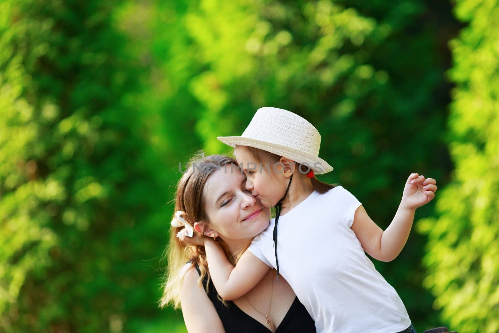 Daughter hugging her mother on the background of bright juicy green foliage. Happy family. Mom and child girl. Space for text. Shallow depth of field. Selective focus.