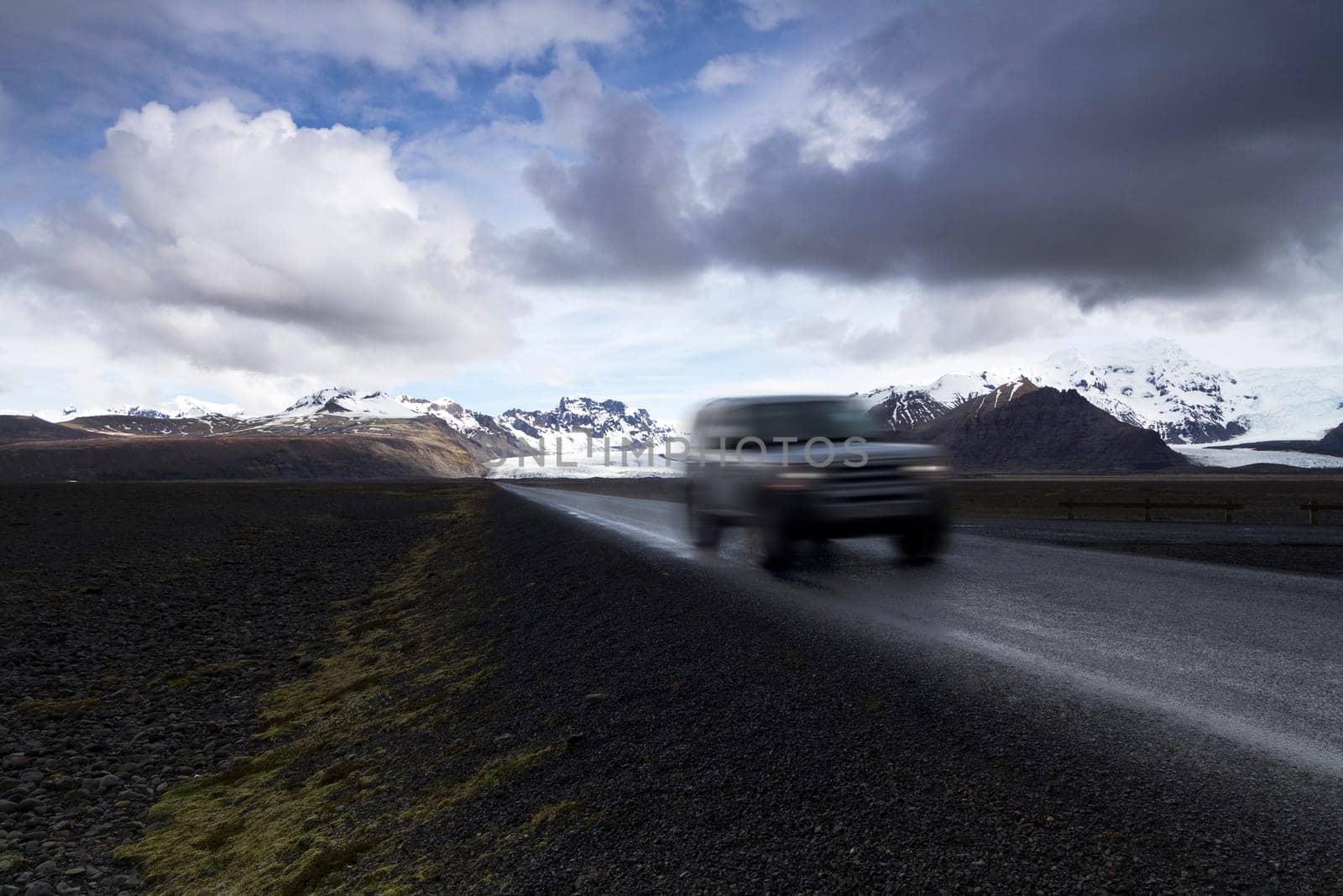 Horizontal photo of an off road car on a straight asphalt road coming from the mountains with clouds above and the Vatnajokull glacier in the background, Iceland