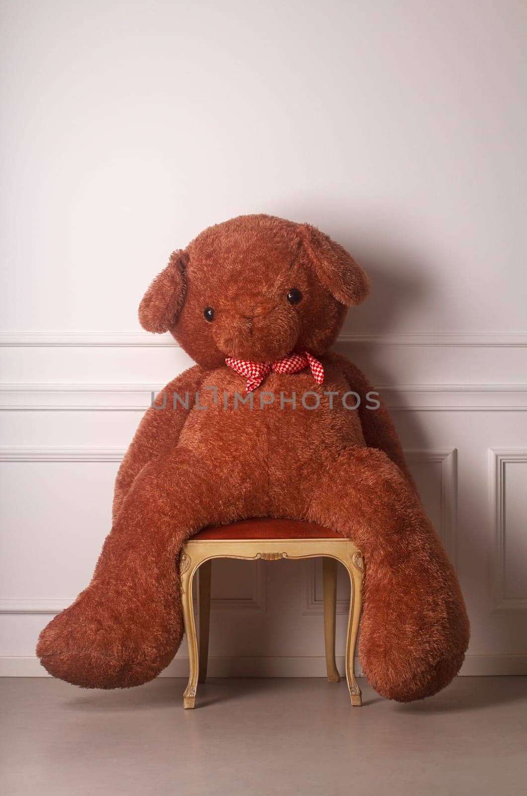 Huge toy bear with a dotted bow standing on a chair against a white wall