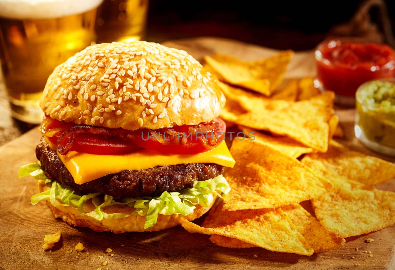 Large sesame seed bun cheeseburger topped with tomatoes, onions and lettuce with nacho chips, salsa, guacamole and beer in background