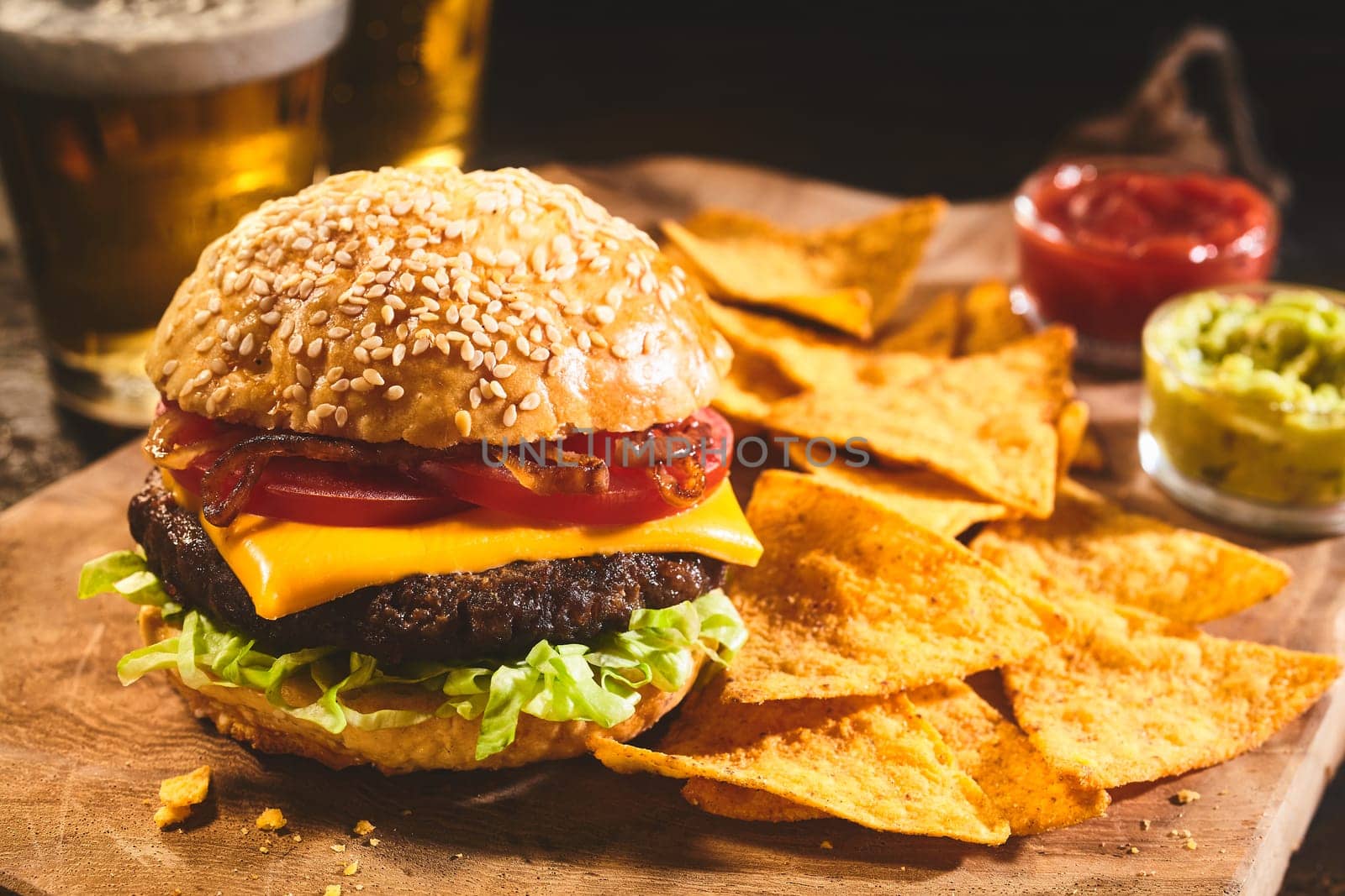 Large freshly prepared cheeseburger topped with tomatoes, onions and lettuce with nacho chips, salsa, guacamole and beer in background