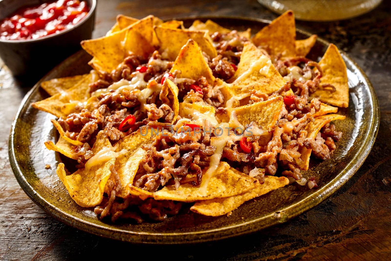 Plate full of yellow corn nachos and cooked ground beef with bowl of red salsa in background over wooden table