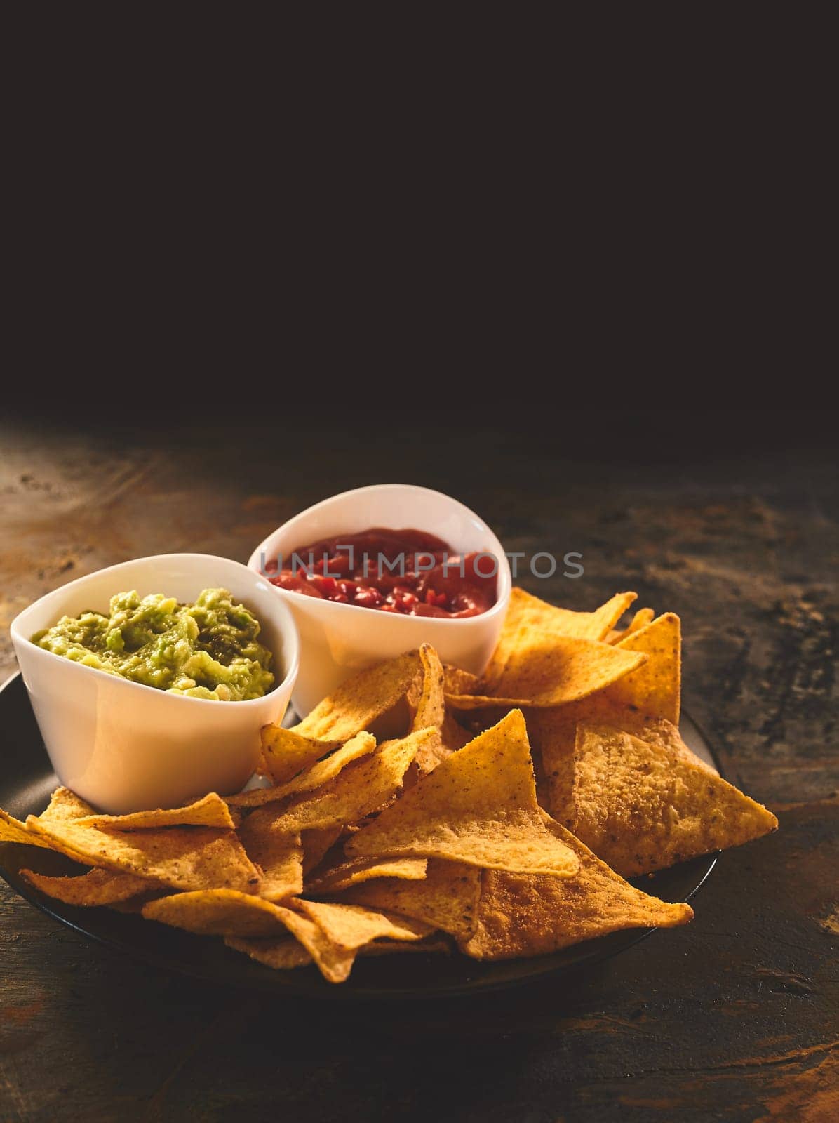 Plate of Mexican cuisine appetizer of triangular yellow corn tortilla chips beside little bowls of guacamole and salsa dip