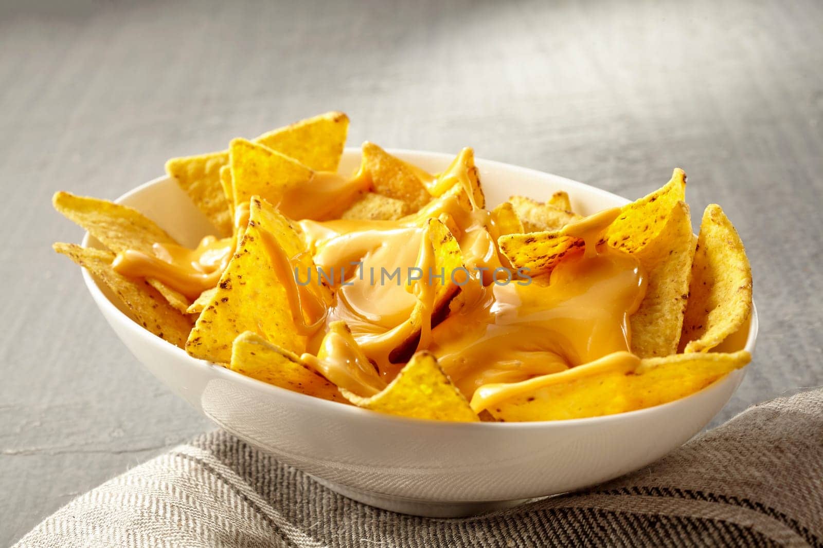 Single oval shaped white bowl of yellow tortilla chips topped with melted cheese placed on folded fabric napkin over tablecloth with copy space