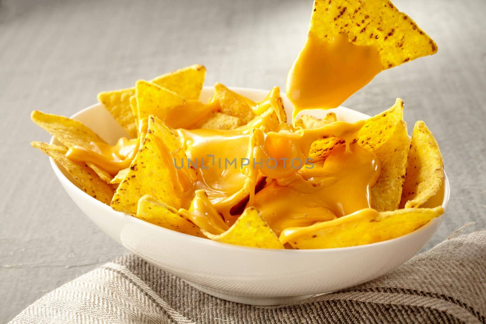 Single triangular yellow corn tortilla chip pulled out of bowl of cheese covered nachos over gray and white tablecloth