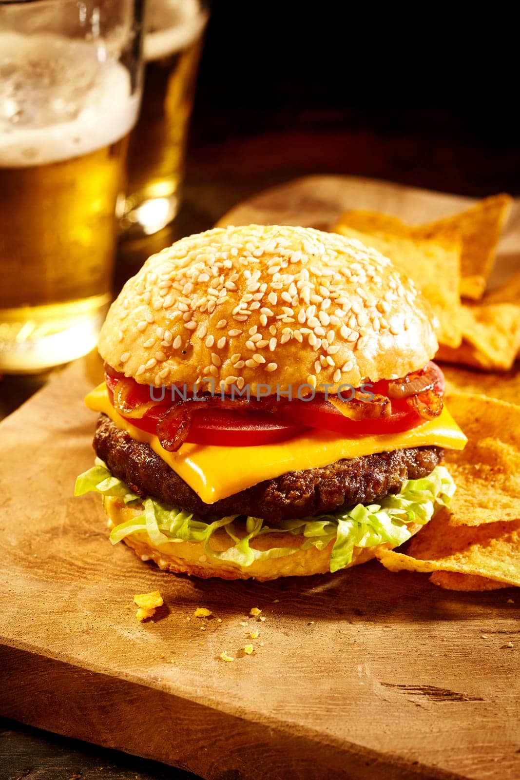 Single delicious well done burger topped with onions, tomatoes, cheddar cheese slice and lettuce with nacho chips on cutting board and two glasses of beer in background