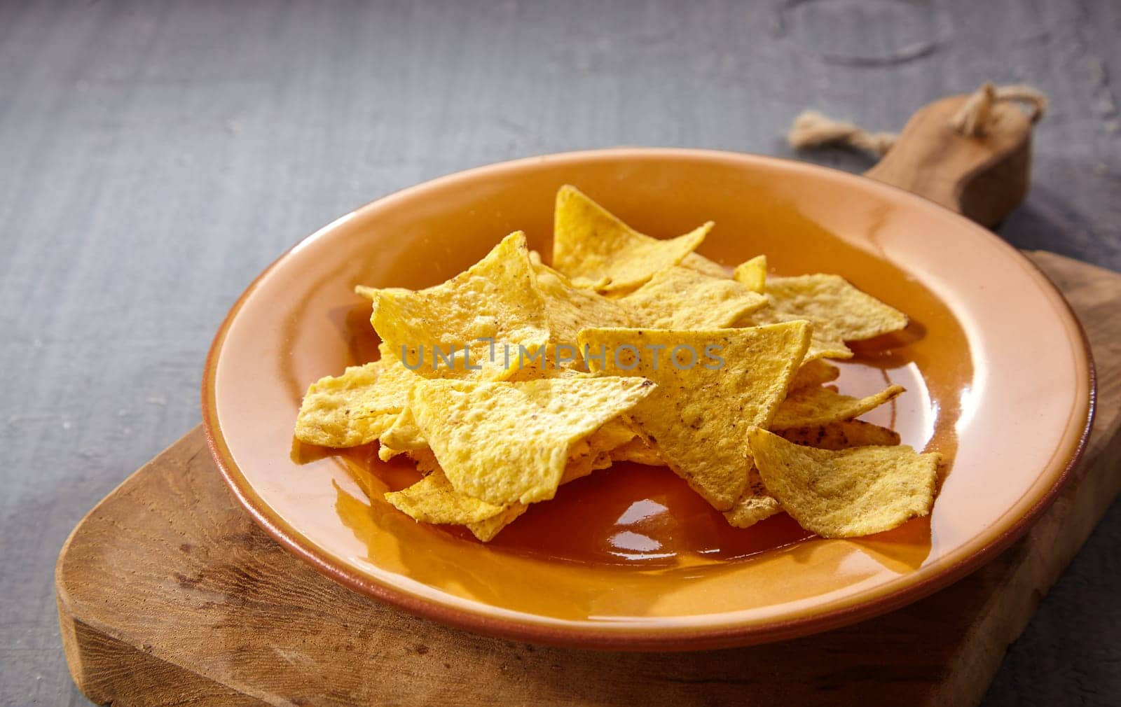 Single plate of tasty yellow tortilla chips on thick wooden cutting board over gray table with copy space