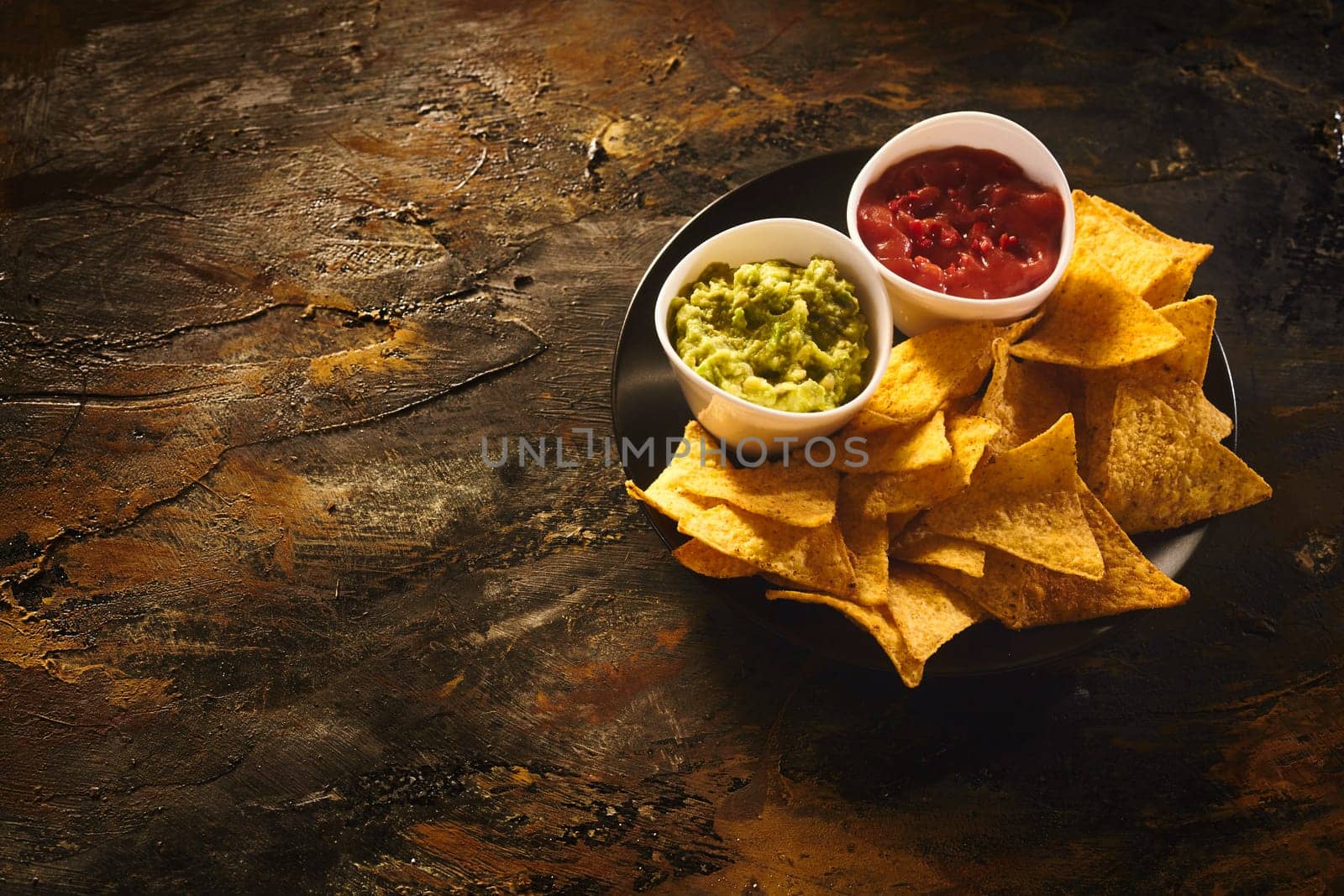 Top down view on delicious crunchy appetizer of triangular yellow corn tortilla chips beside little bowls of guacamole and salsa dip over old dark wooden table with copy space