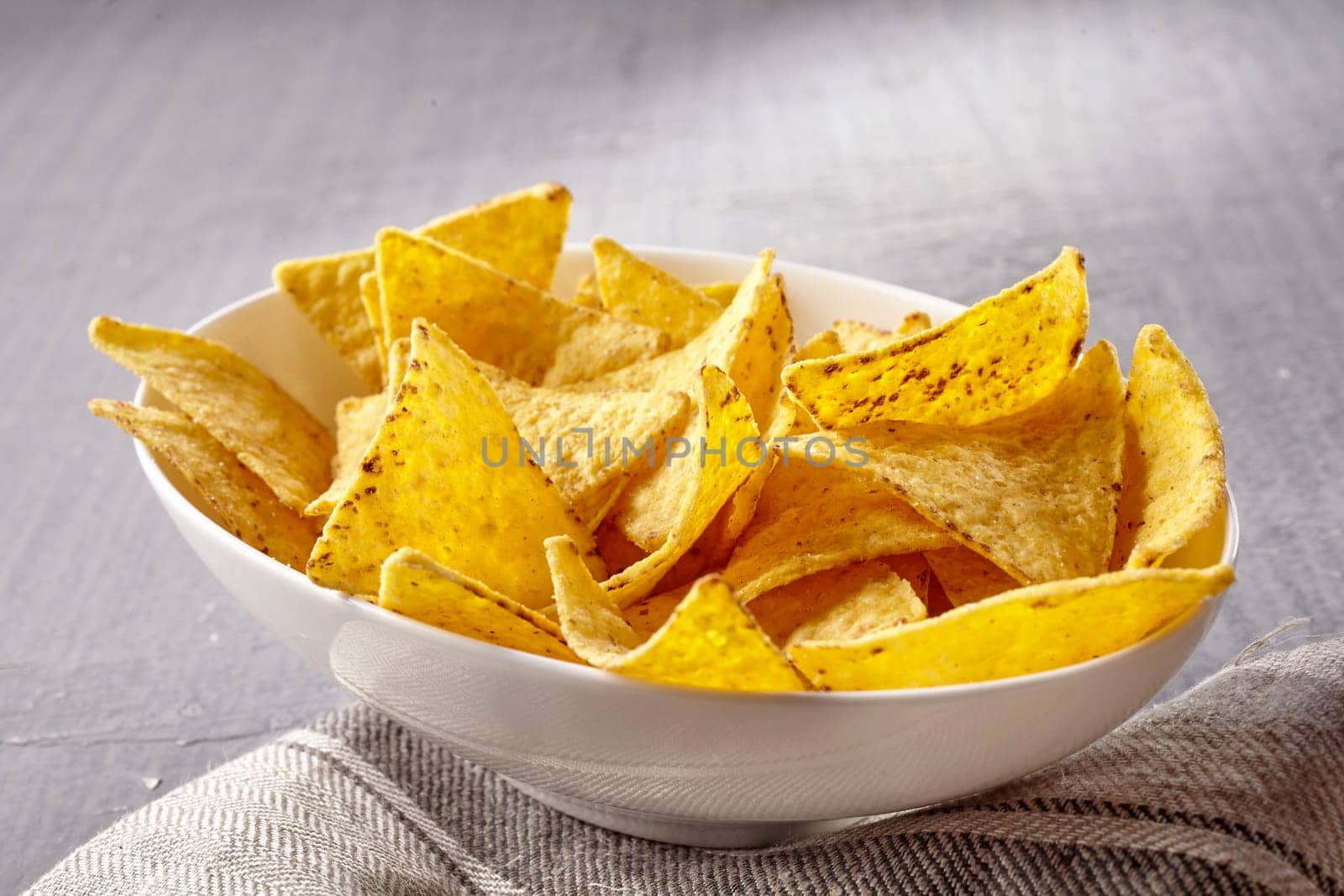 Single oval shaped white bowl of yellow tortilla chips placed neatly on folded fabric napkin over tablecloth with copy space