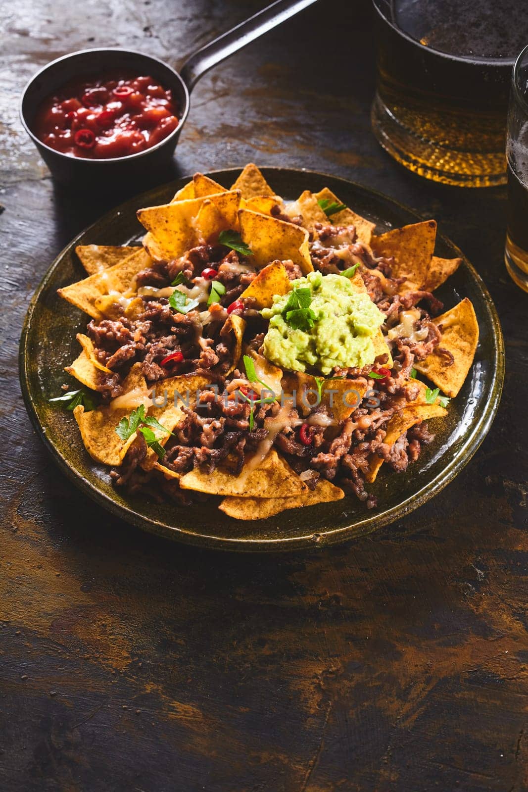 Red salsa hot sauce in bowl beside large ceramic plate of yellow corn tortilla chips, guacamole and beer on dark wooden table with copy space
